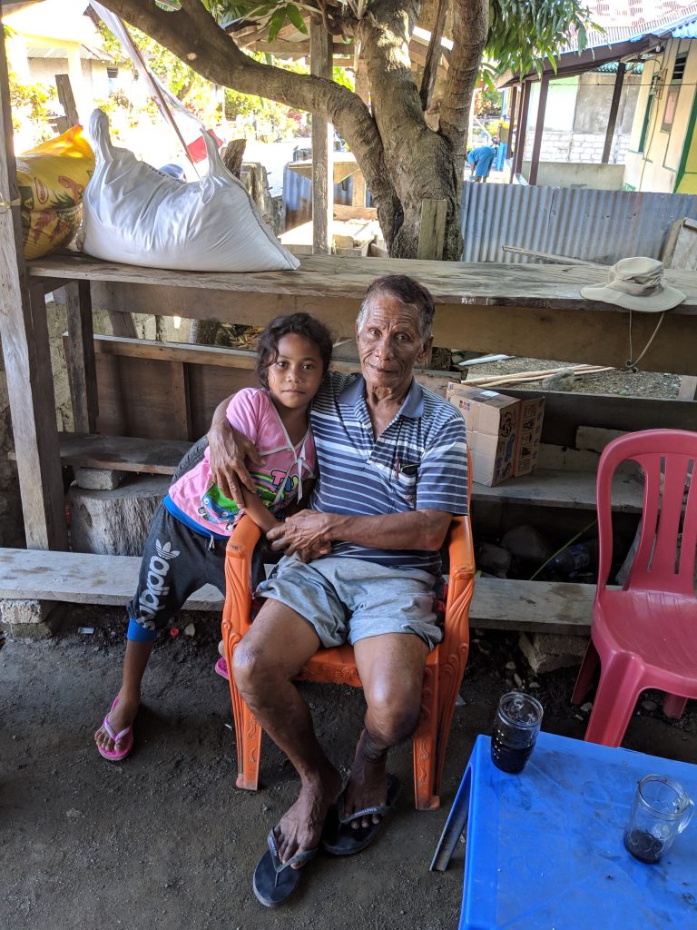 Man sitting outdoors with a child in the shade of a tree next to a house.