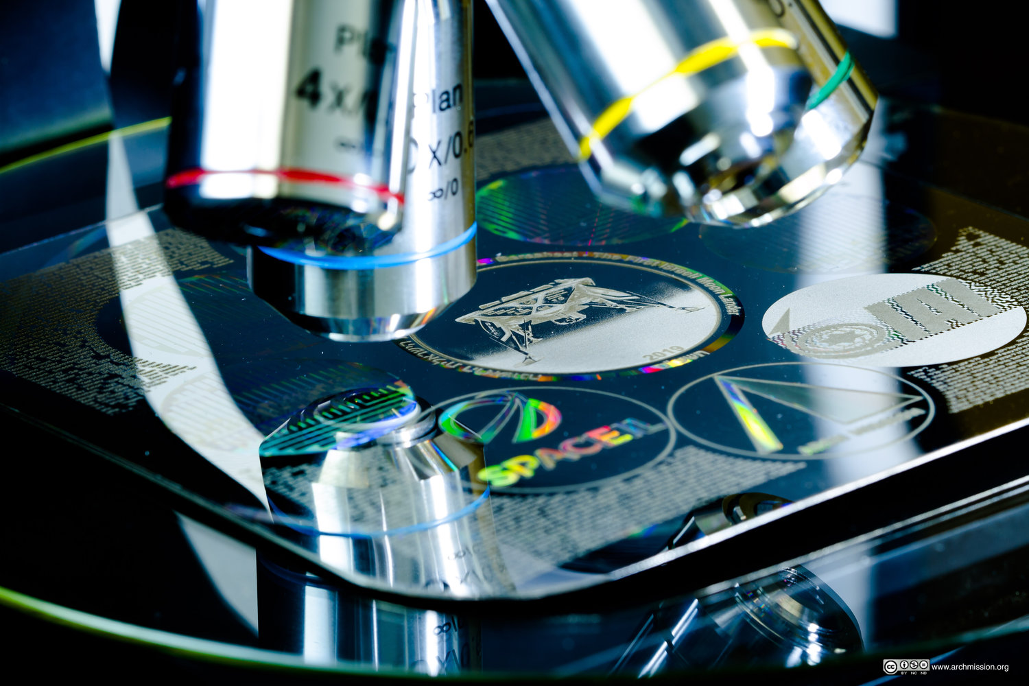 Detail of shiny nickel disc with SpaceIL icons and blurred microscopic "pages" under microscope lenses. 