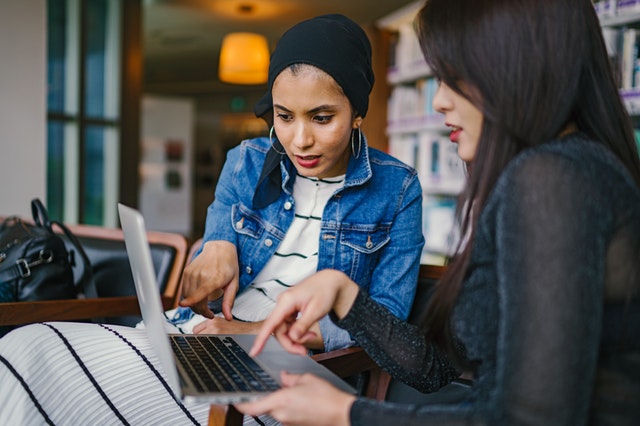 Two women are sitting in an office or library looking at a laptop screen.